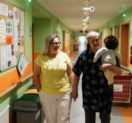 Two women walking in a nursing home corridor, one holding a child doll.