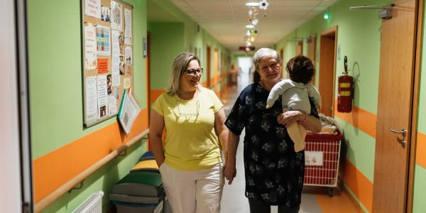 Two women walking in a nursing home corridor, one holding a child doll.